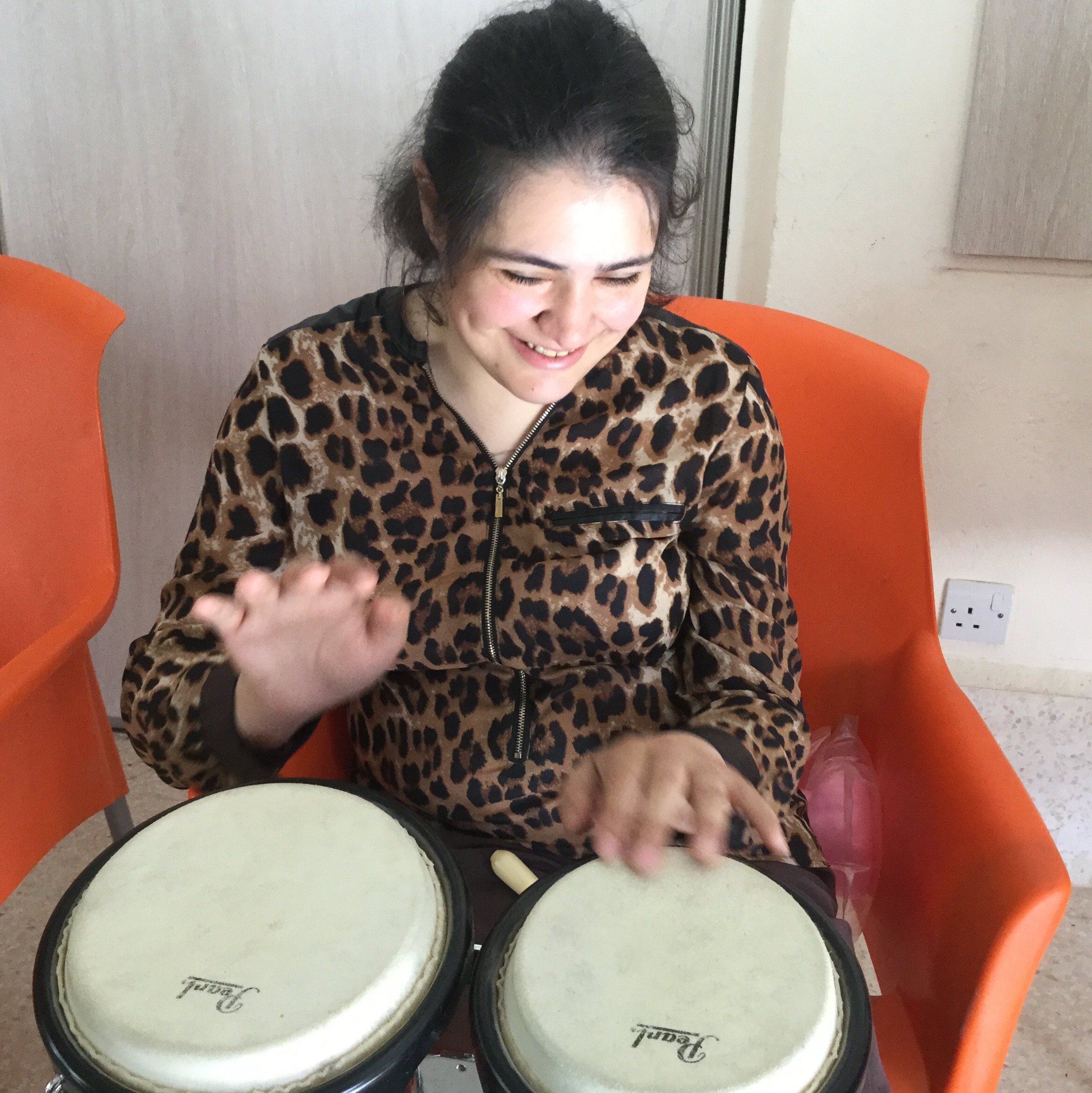 Young woman with dark hair smiling as she looks down at the hand drums she is playing. She is wearing a leopard print top and is sat on an orange chair.