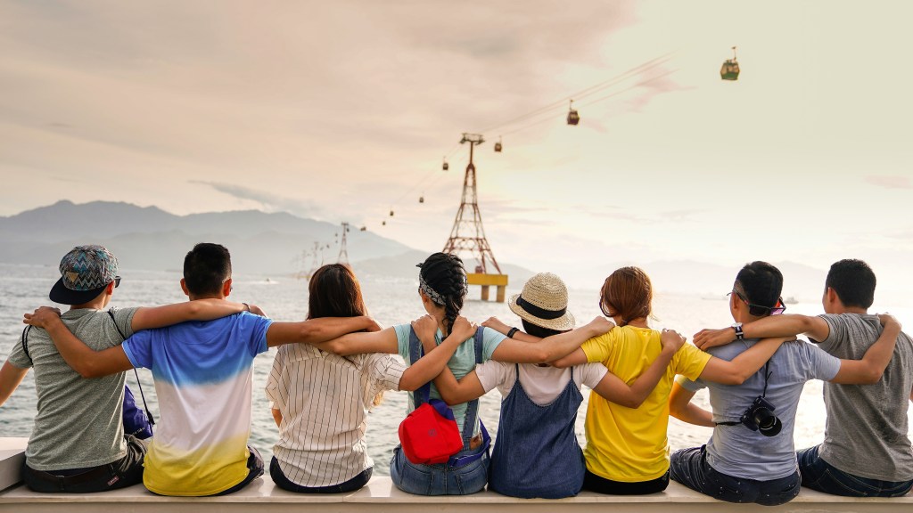 Eight people sat on a sea wall looking out at the water. They have their arms around each others' shoulders. In the distance are mountains. A skyrail runs over the water, supported by pylons.